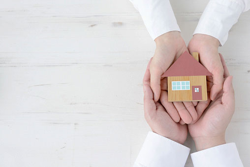 A wooden toy house being held in a set of hands, and those hands are being held by a larger set of hands, to illustrate the value of emotional support during divorce-related real estate sales