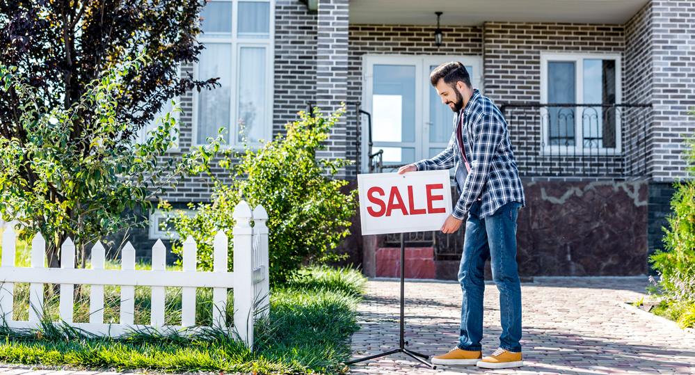 Photo of a home, a man is placing a For Sale sign out front